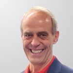 Smiling man in a navy blazer and red collared shirt against a light gray background. His expression is friendly and welcoming.