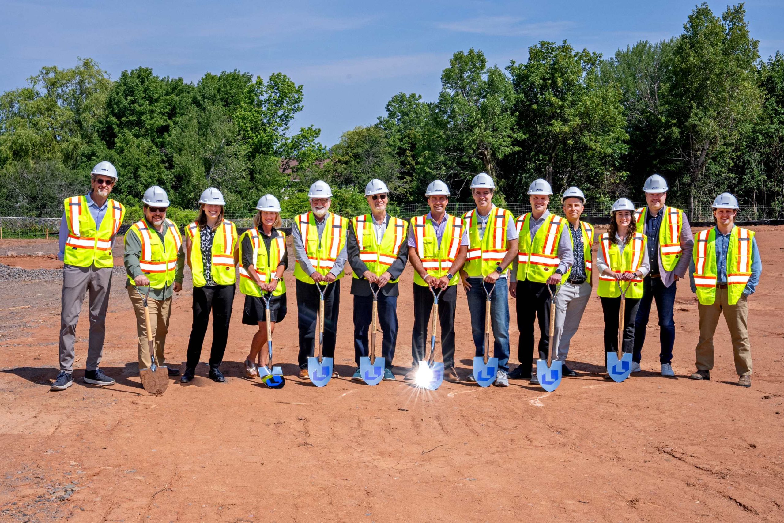 A group of thirteen people in safety vests and hard hats poses with shovels on a construction site under a clear blue sky.