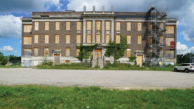 A large, abandoned brick building with boarded-up windows and overgrown ivy. Scaffolding is on the right side. The sky is partly cloudy, and there's a grassy area in the foreground.