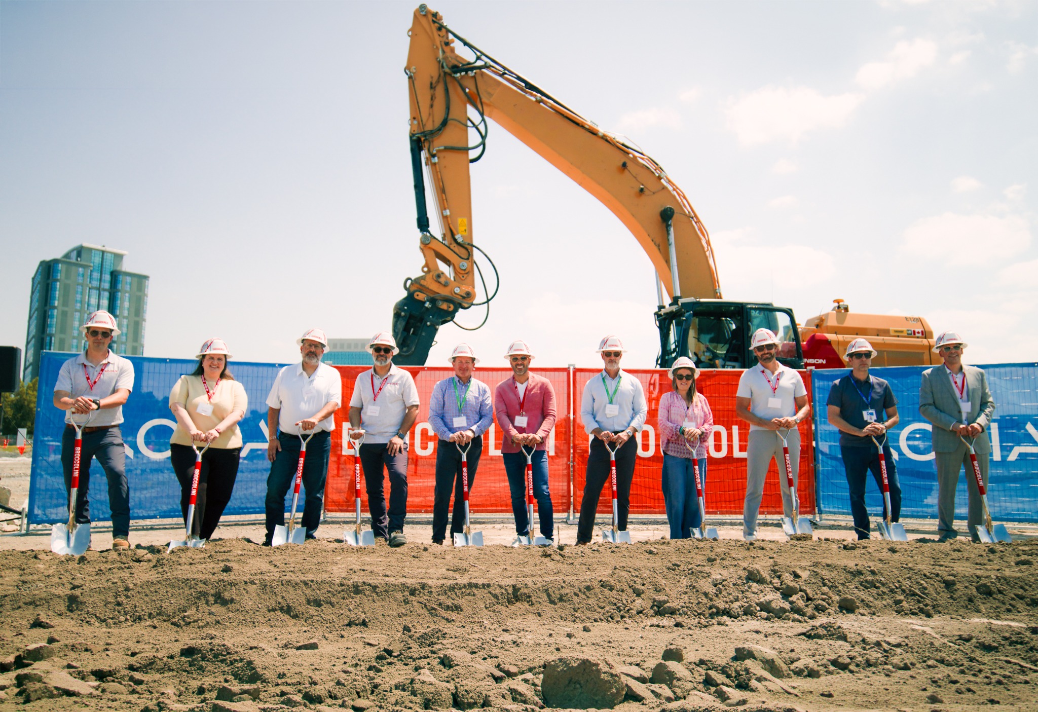 A group of ten people in hard hats and business attire stand with shovels at a construction site. An excavator is in the background under a clear sky.