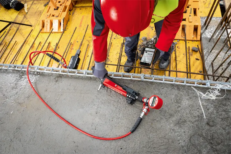 A construction worker in red safety gear uses a power tool on a concrete surface at a building site, with equipment and rebar visible nearby.