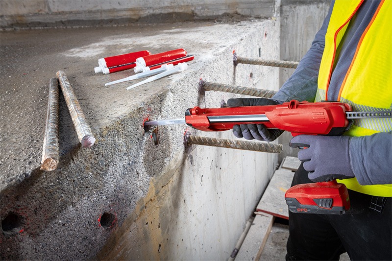 A construction worker uses a red caulking gun to apply adhesive into holes in a concrete block, with steel rods and adhesive tubes nearby.