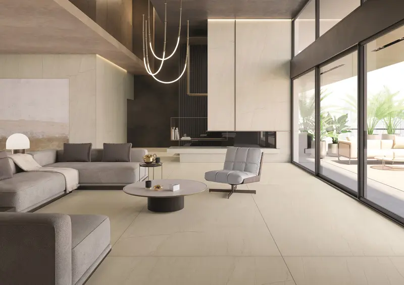 Modern living room with gray sectional sofa, stylish chairs, and round coffee tables. Large windows reveal a bright patio with greenery.