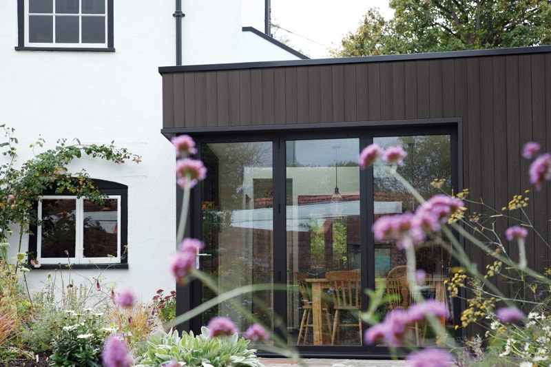 A modern house with a white exterior and dark brown extension featuring large glass doors, surrounded by colorful flowers and greenery.
