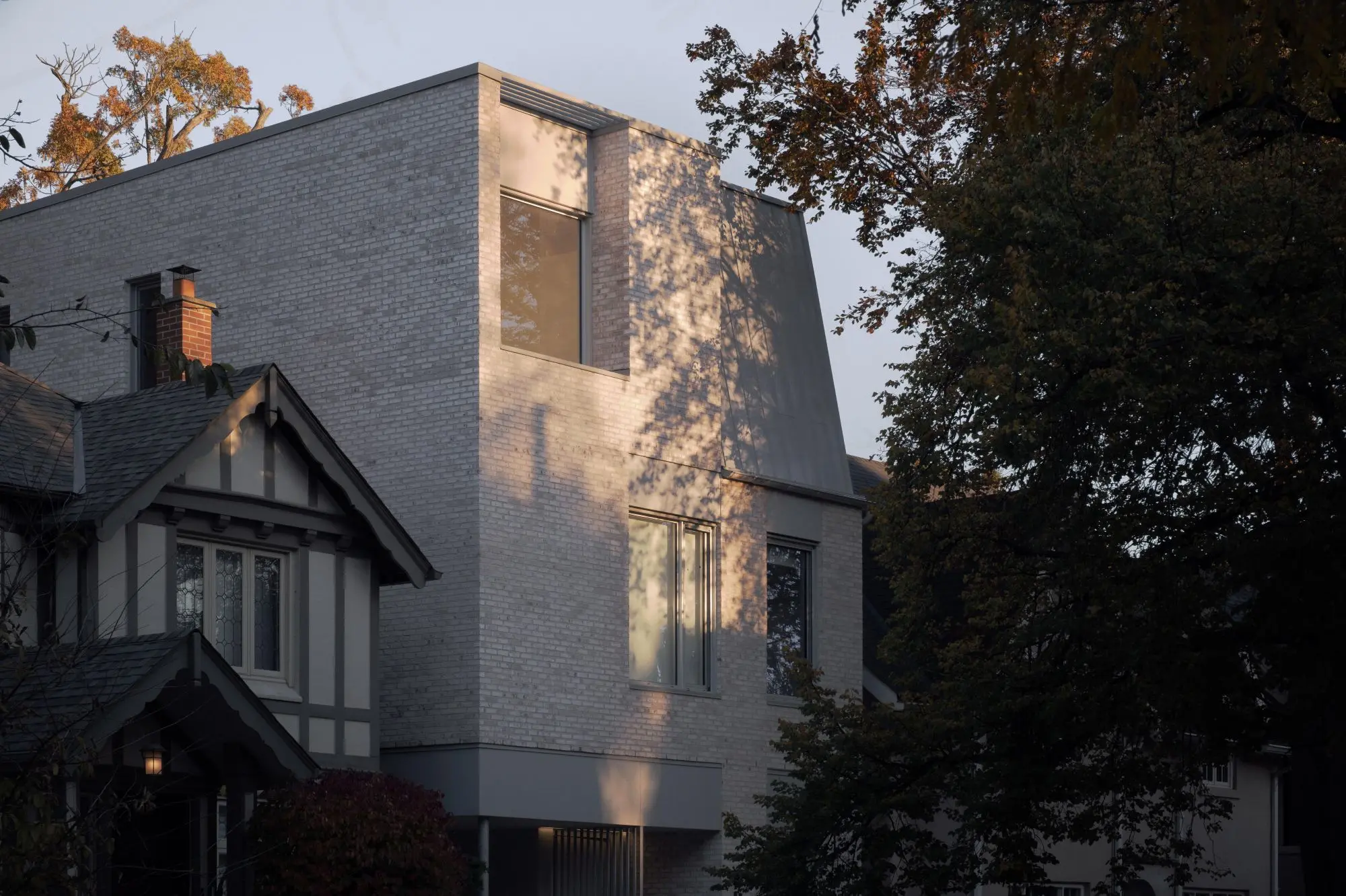 A modern light-brick building with large windows stands beside an older, traditional house, framed by autumn-colored trees.