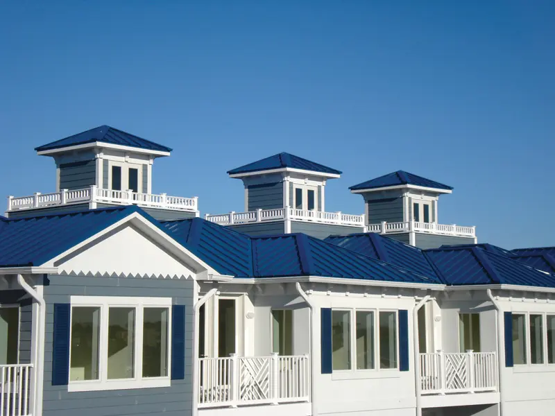 Row of three coastal-style buildings with blue metal roofs and white exteriors, featuring large windows and balcony railings against a clear blue sky.