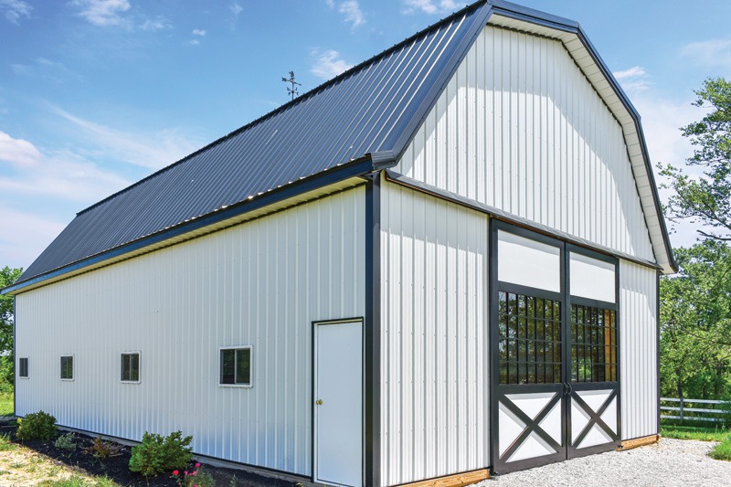 A modern white barn with a dark metallic roof, featuring small windows and a double door, set in a rural landscape with greenery.