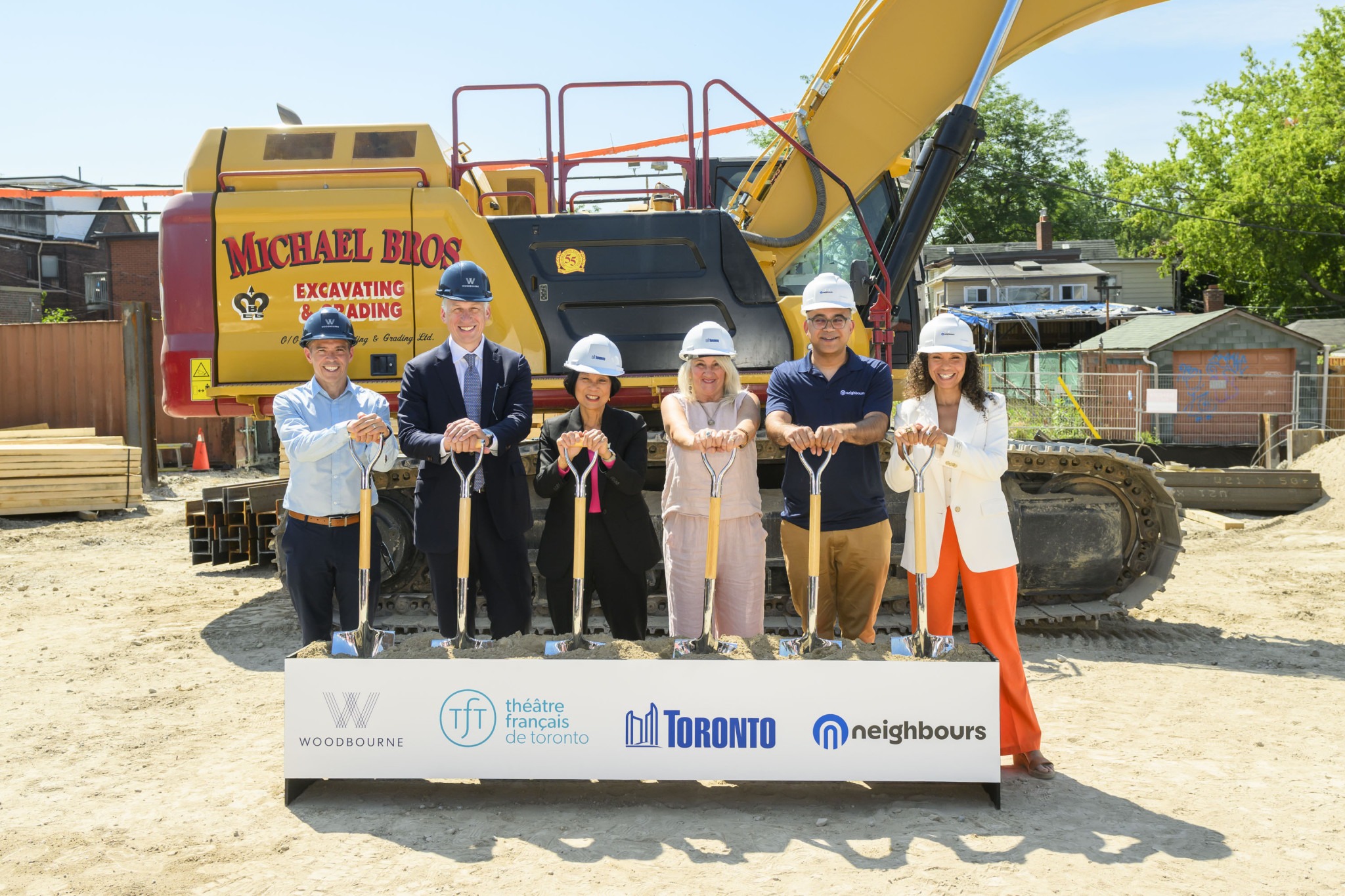Group of six people in hard hats participating in a groundbreaking ceremony, holding shovels in front of a yellow excavator. A banner with logos is displayed in front.