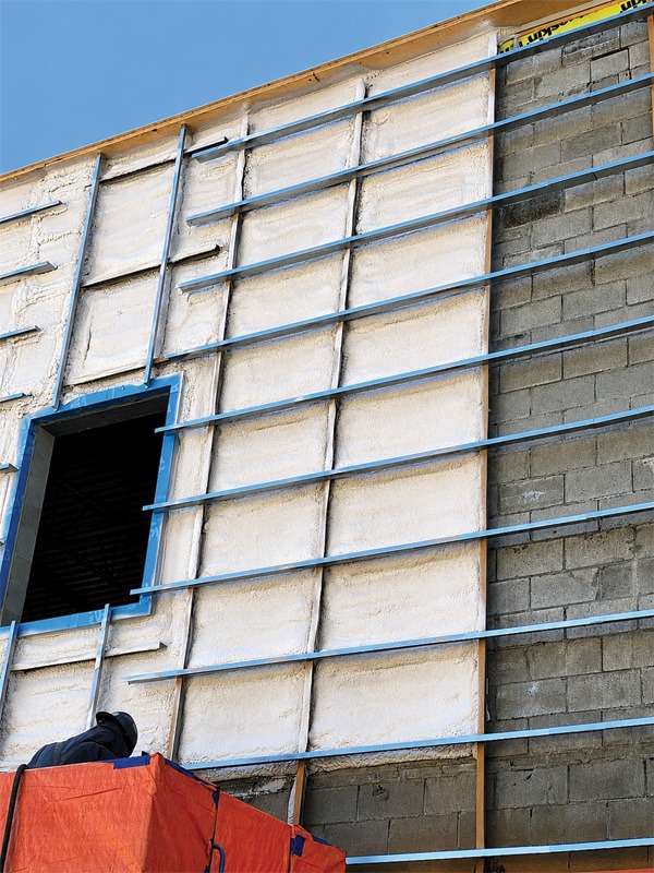 Close-up of a construction wall showing a mix of stucco and concrete, with blue support bars, an open window, and a worker on a red-covered platform.