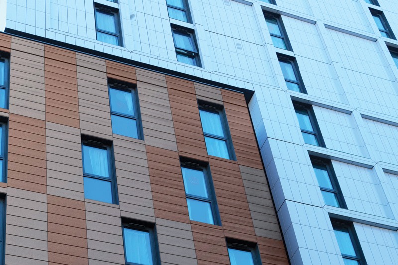 A building façade with two sections: the left in varying shades of brown with dark-framed windows, and the right in a uniform white design with thin black window frames.