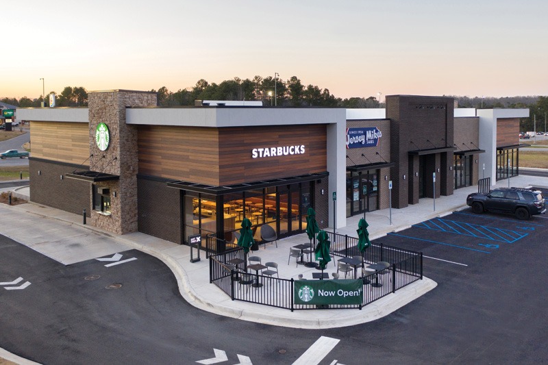 A modern Starbucks building with a stone and wood facade, outdoor seating under green umbrellas, and a "Now Open!" banner, next to a Jersey Mike's restaurant.