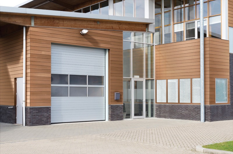 Modern building entrance featuring a large gray garage door, glass doors, and wooden paneling, with a paved area and grassy patch.