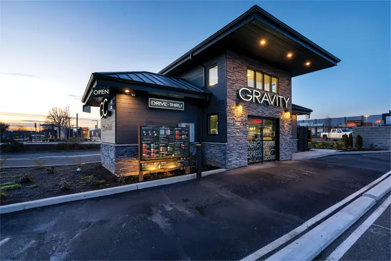 A modern drive-thru restaurant named "GRAVITY" with a dark wood and stone exterior, illuminated signage, and a menu board, set against a twilight sky.