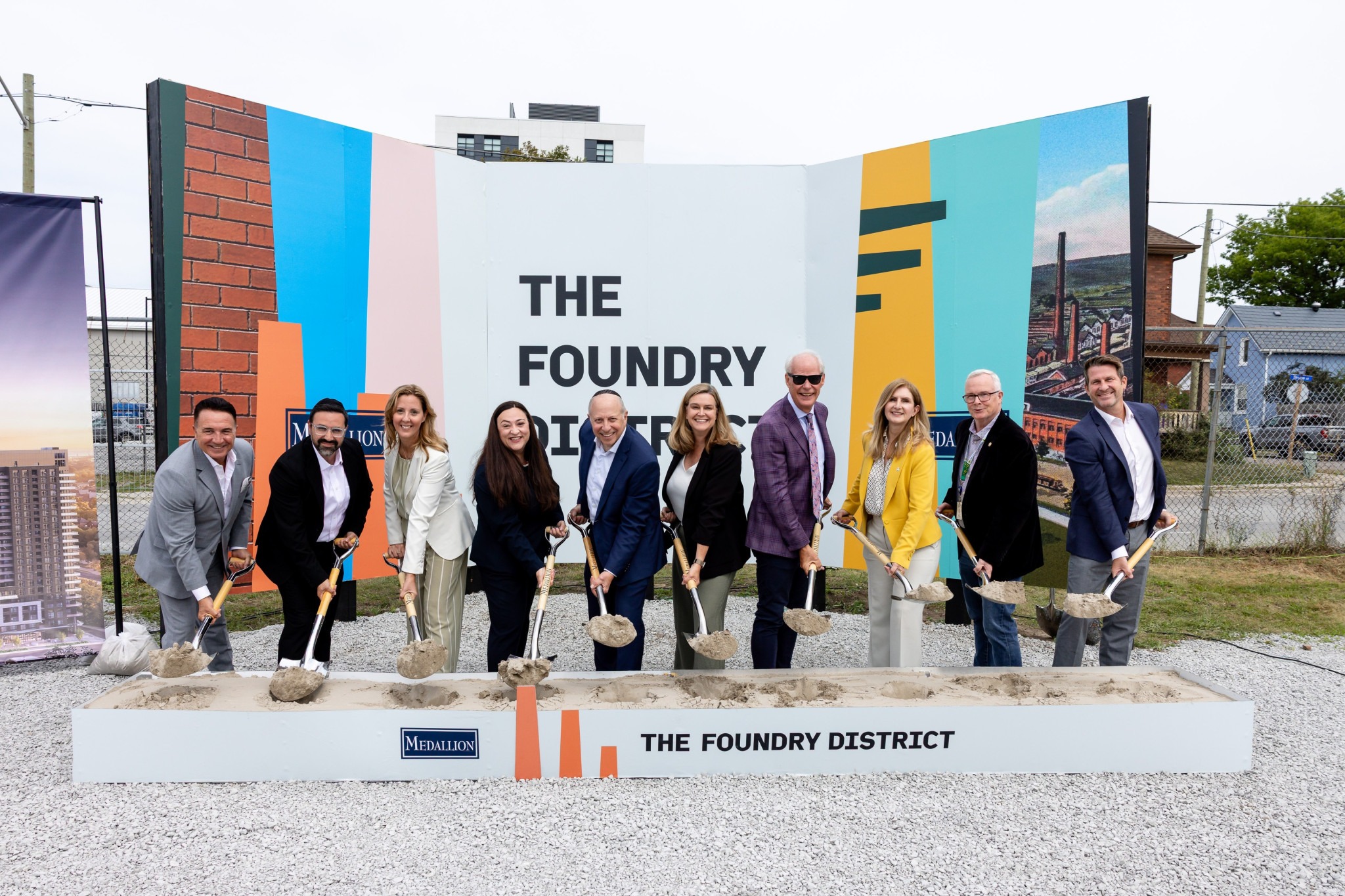 A group of ten individuals holds shovels at a groundbreaking ceremony for "The Foundry District," standing in front of a colorful backdrop displaying the project's name.