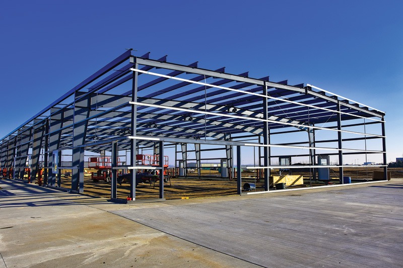 A steel framework of a building under construction with scissor lifts and materials on gray concrete flooring under a clear blue sky.
