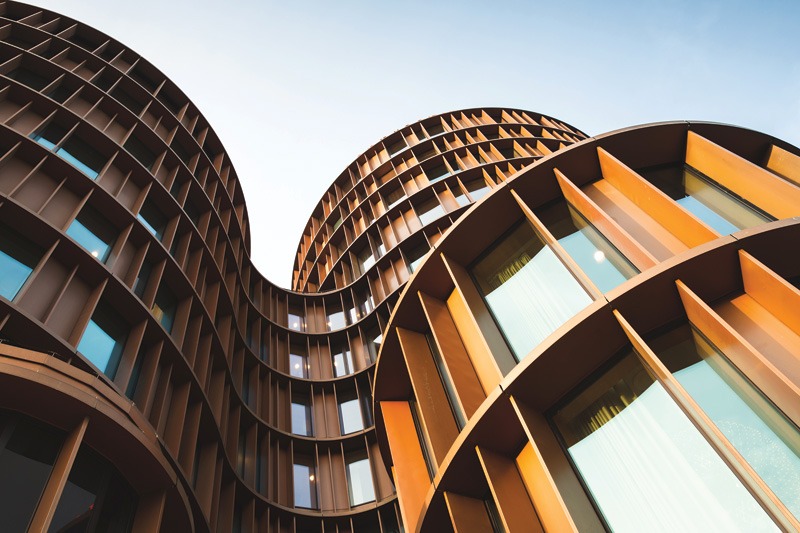 A modern building with curved, reddish-brown facades and large rectangular windows, set against a clear blue sky.