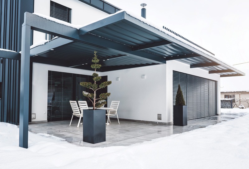 Modern outdoor patio with a flat-roofed structure, featuring a gray tiled floor, white metal chairs, and evergreen plants in black planters, all set against a snowy background.