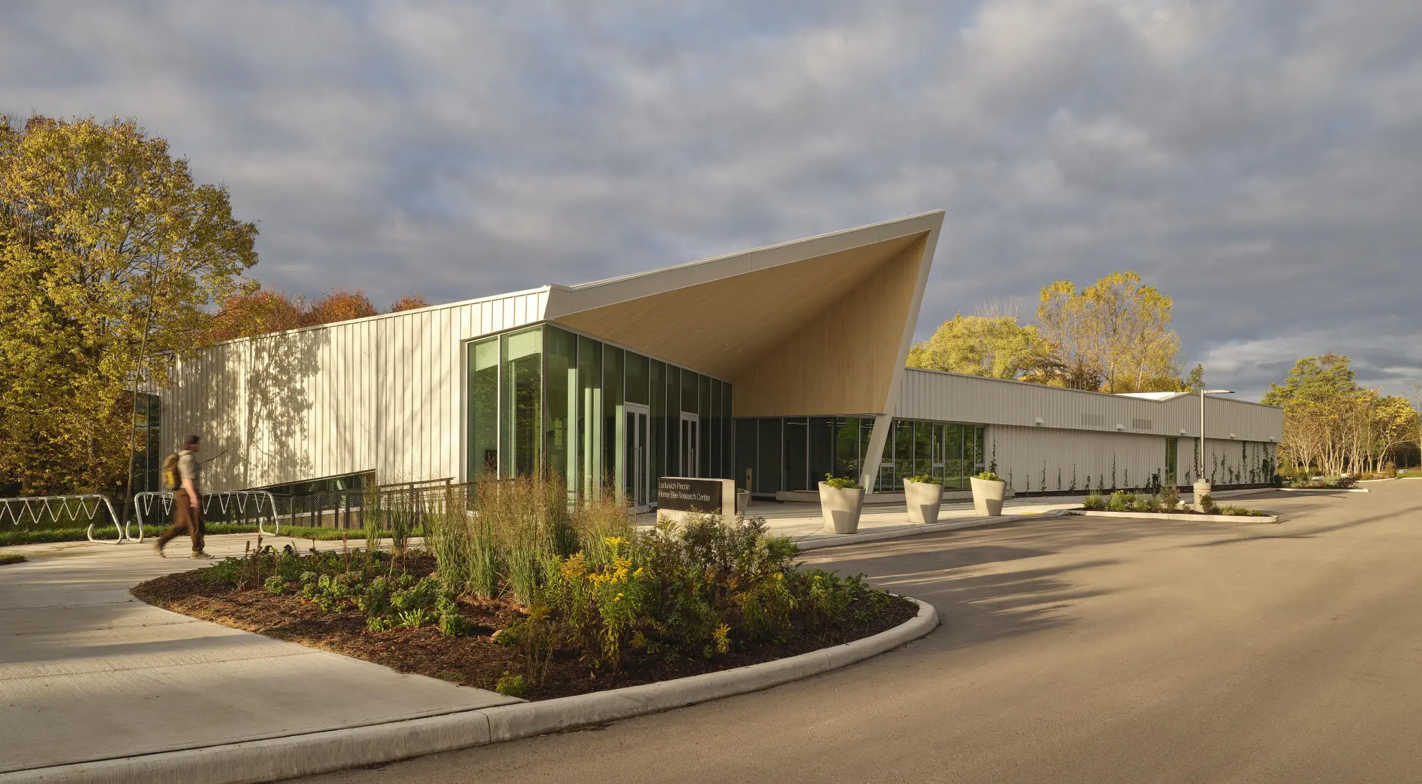 Modern research center with a peaked roof and large glass facade, surrounded by landscaped greenery and a pathway. A person walks along the path in the foreground.