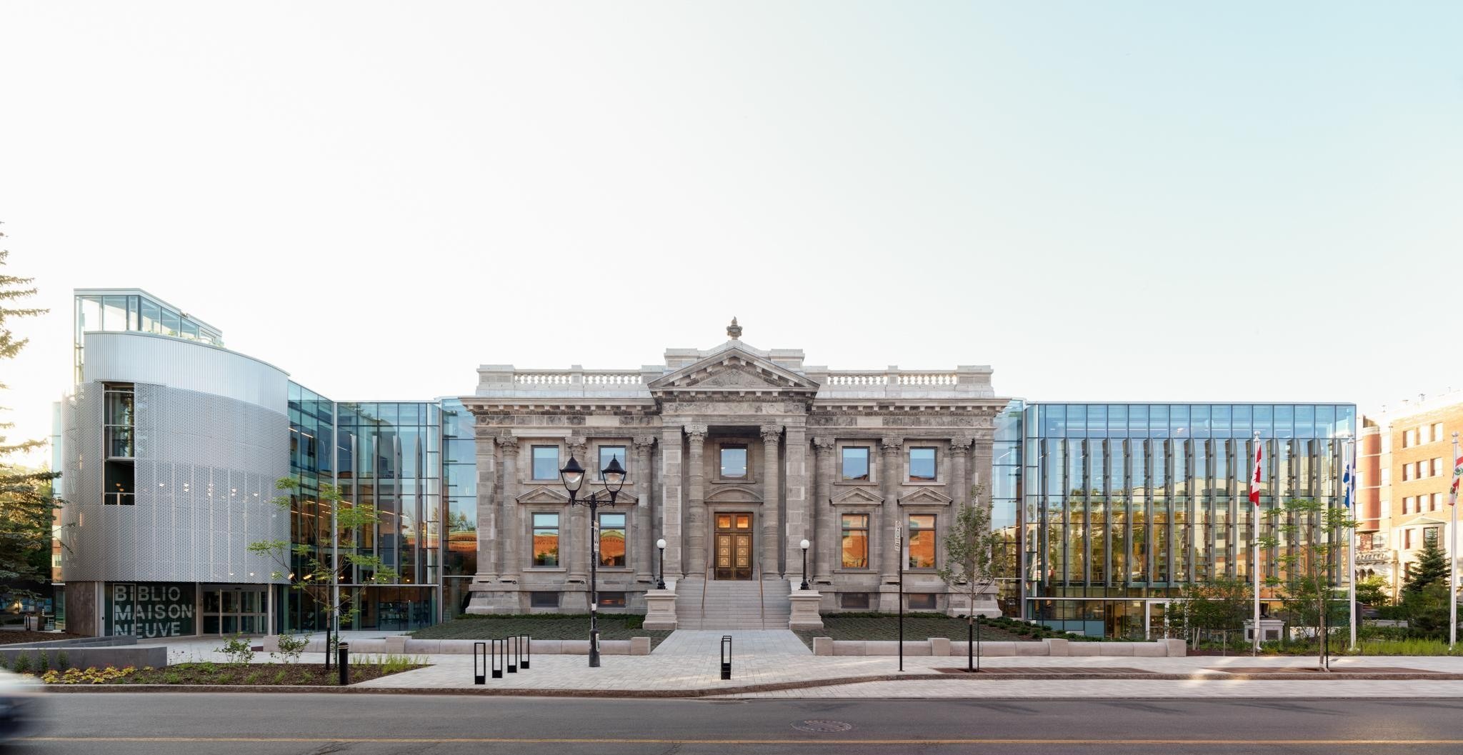 A juxtaposition of a classical stone building with ornate details beside a modern glass and metal structure, set against a clear sky, featuring landscaped greenery in the foreground.