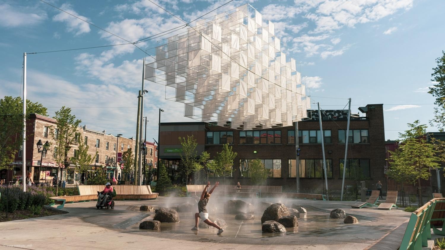 An urban park features a transparent suspended installation above a fountain with a child dancing in the water. A person in a wheelchair is seen on a path, surrounded by shops and greenery under a blue sky.