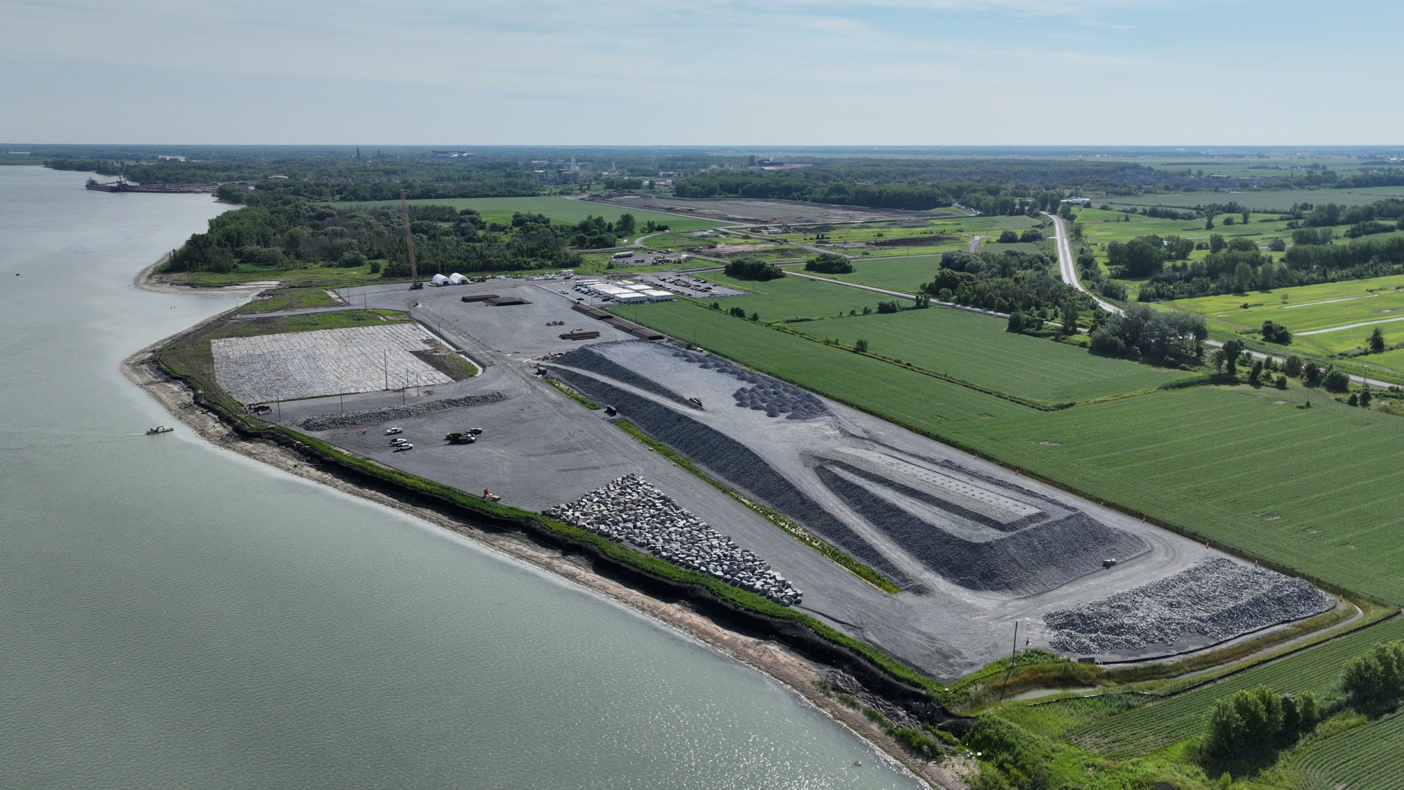 Aerial view of a gravel industrial area next to calm water and green fields, with vehicles and structures visible.