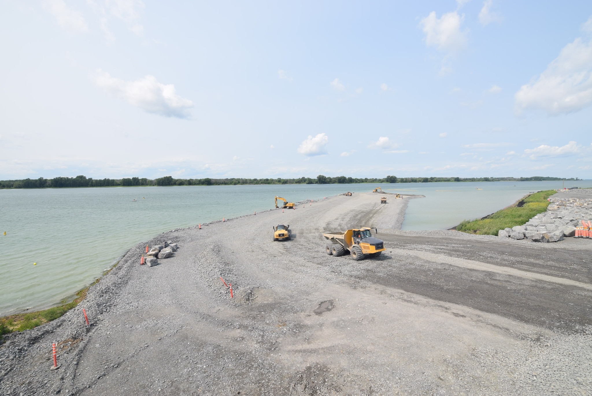 Construction site with yellow trucks and excavator on gravel by calm greenish water, surrounded by trees and blue sky.