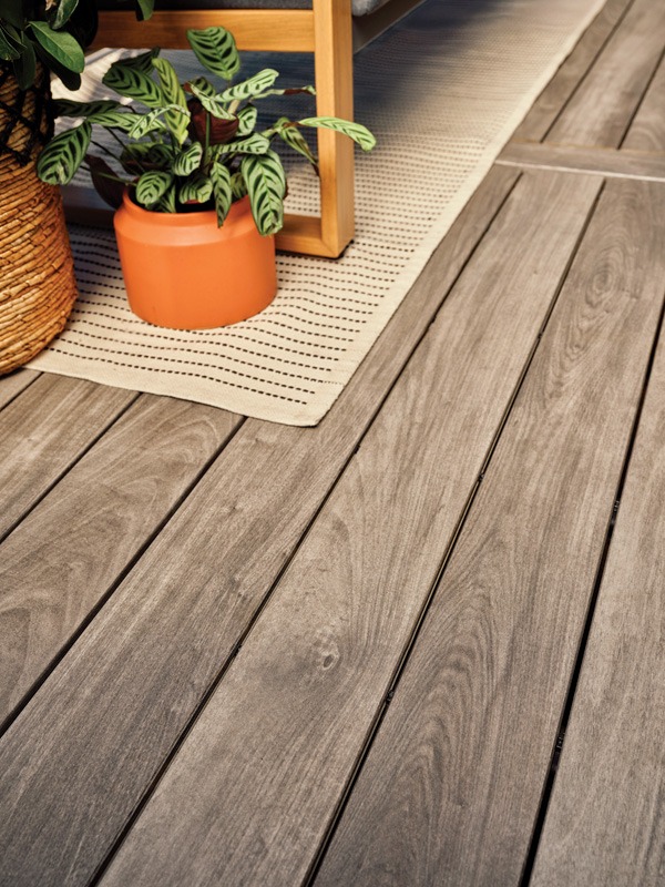 A close-up of a wooden deck with light gray planks, featuring a textured beige rug, an orange pot with a green plant, and wooden furniture in the background.