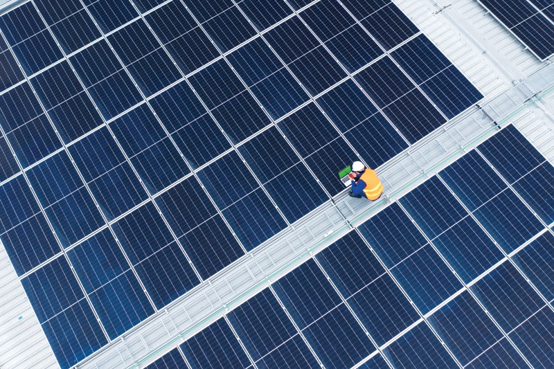 A worker in a yellow safety vest and white hard hat sits on solar panels on a rooftop, holding a green tool.