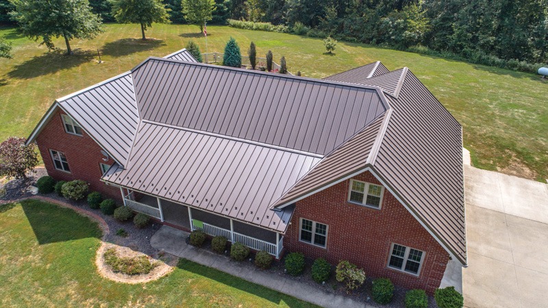 Aerial view of a house with a dark brown metal roof and red brick walls, surrounded by a well-kept yard and greenery.