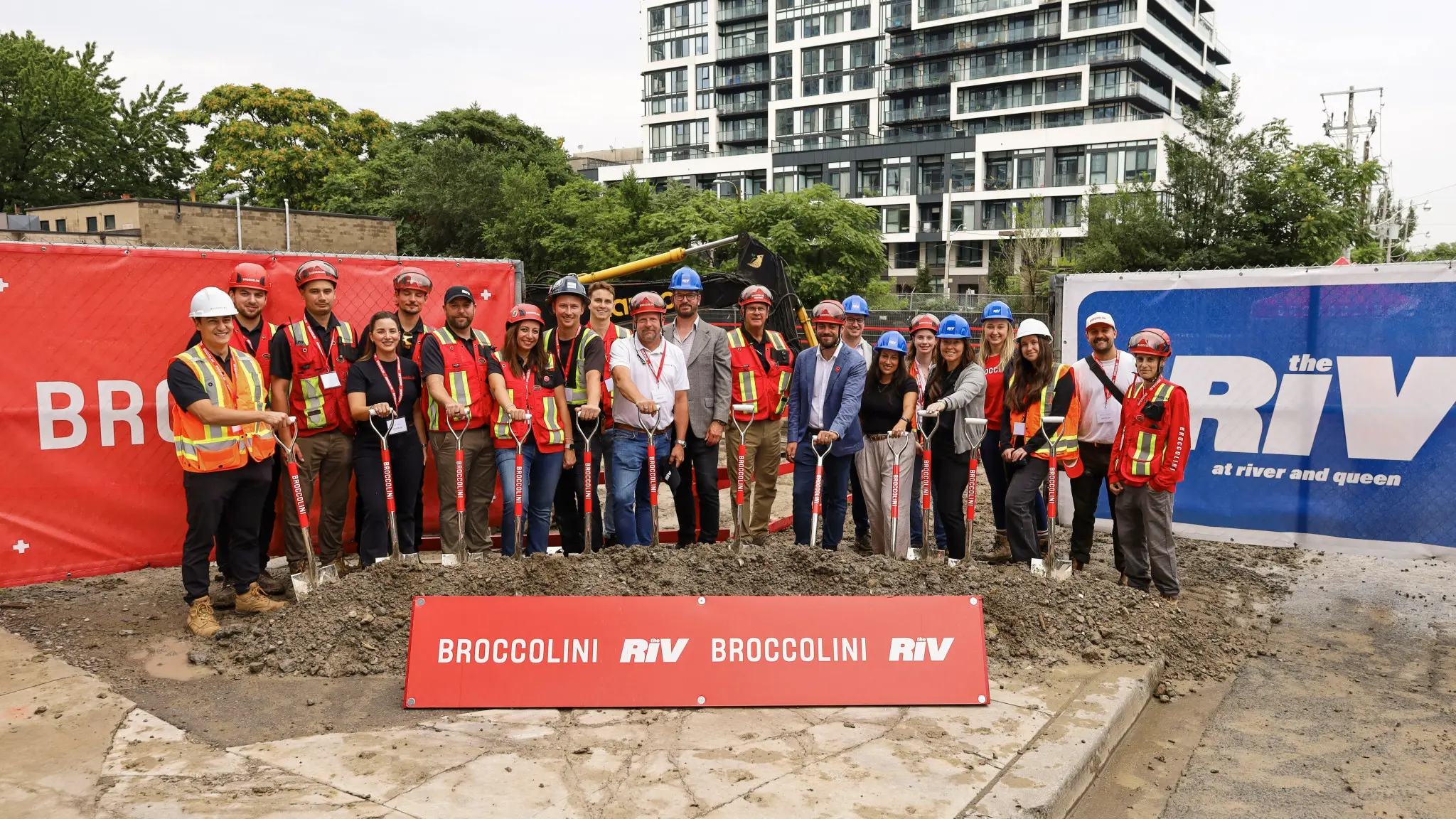 A group of individuals in safety gear celebrates a groundbreaking ceremony at an urban construction site, standing in front of a mound of dirt and holding shovels.