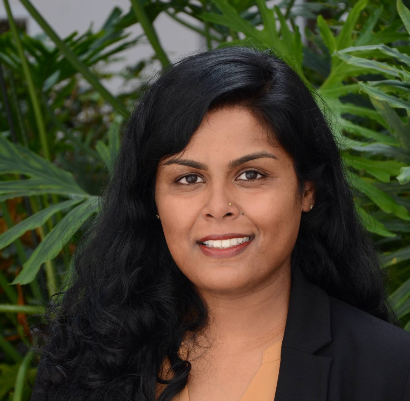 Smiling woman with long black hair wearing a black blazer and peach blouse, photographed in front of green foliage.