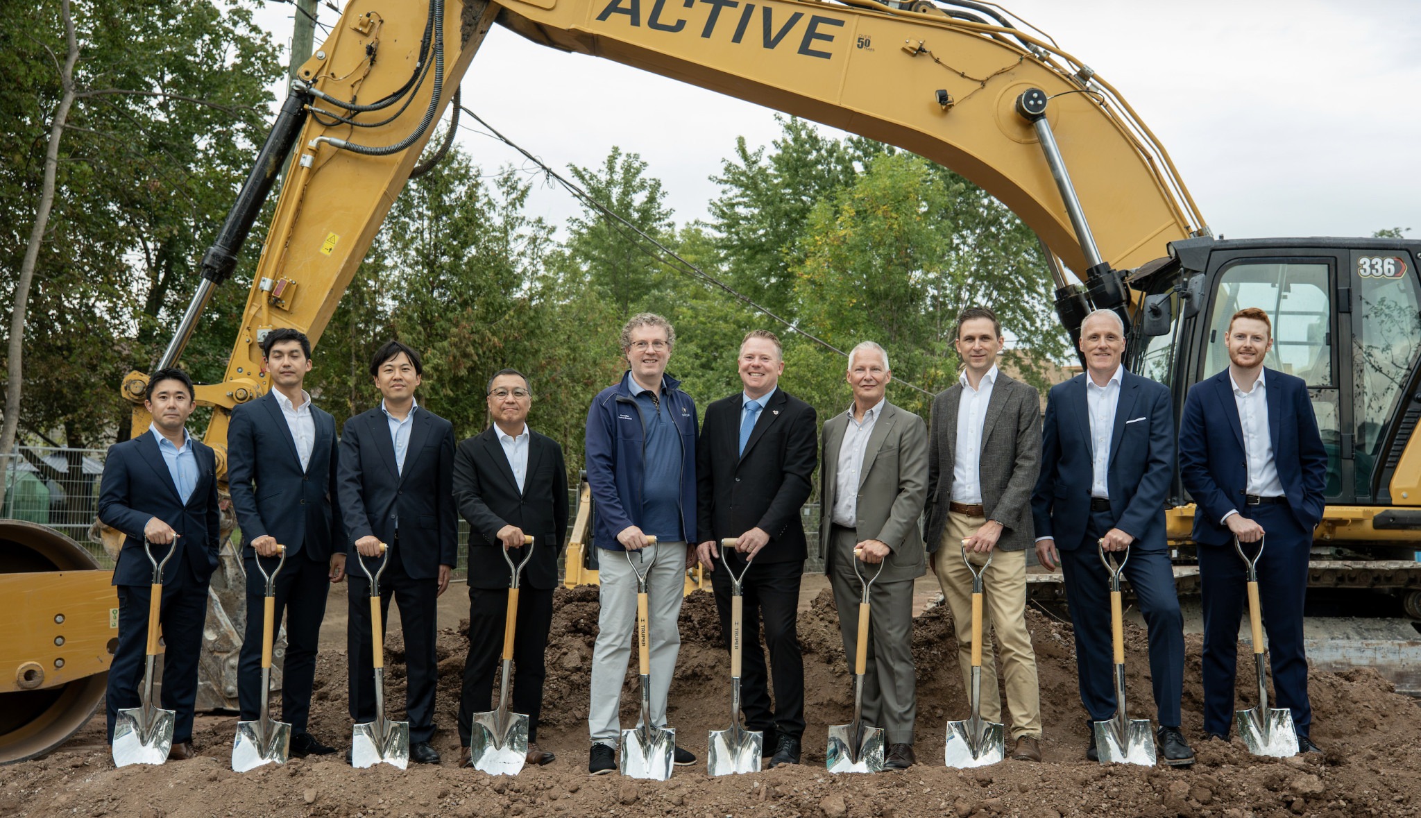 Ten adults pose with ceremonial shovels in front of a large yellow excavator at a groundbreaking ceremony.