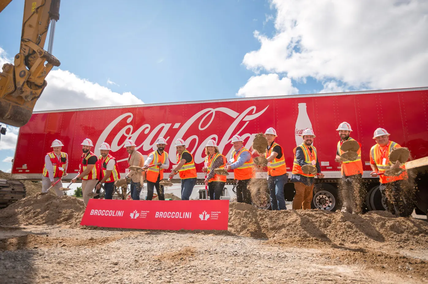 Construction workers toss soil with shovels at a groundbreaking ceremony in front of a red Coca-Cola trailer and a Broccolini sign.