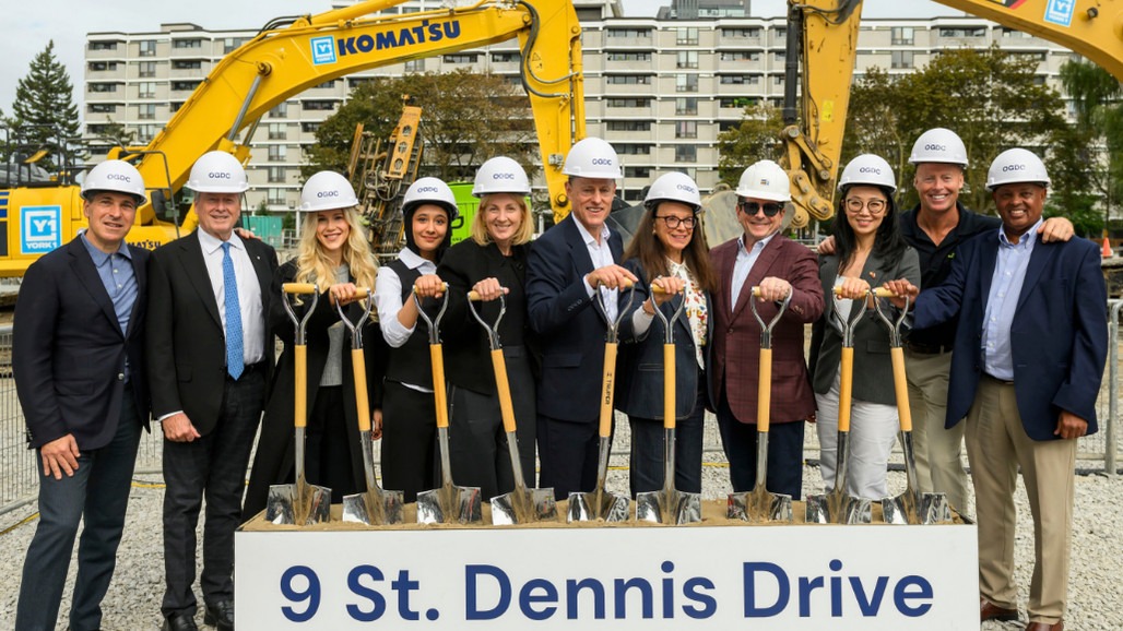 Eleven adults in hard hats hold ceremonial shovels at a construction site in front of yellow excavators and a “9 St. Dennis Drive” sign.