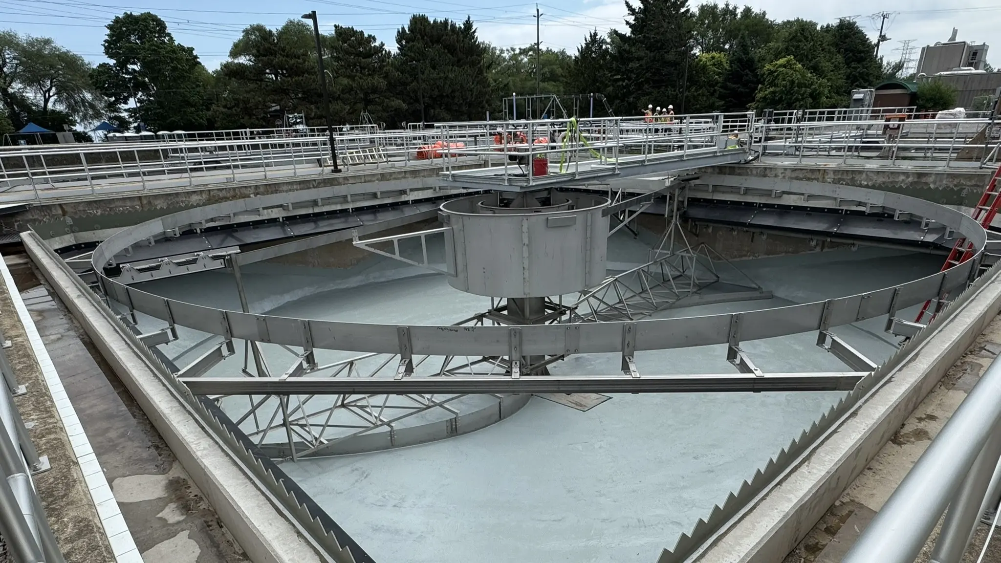 An empty circular industrial treatment basin with a central mechanical scraper, surrounding walkways, railings, and workers in the background.