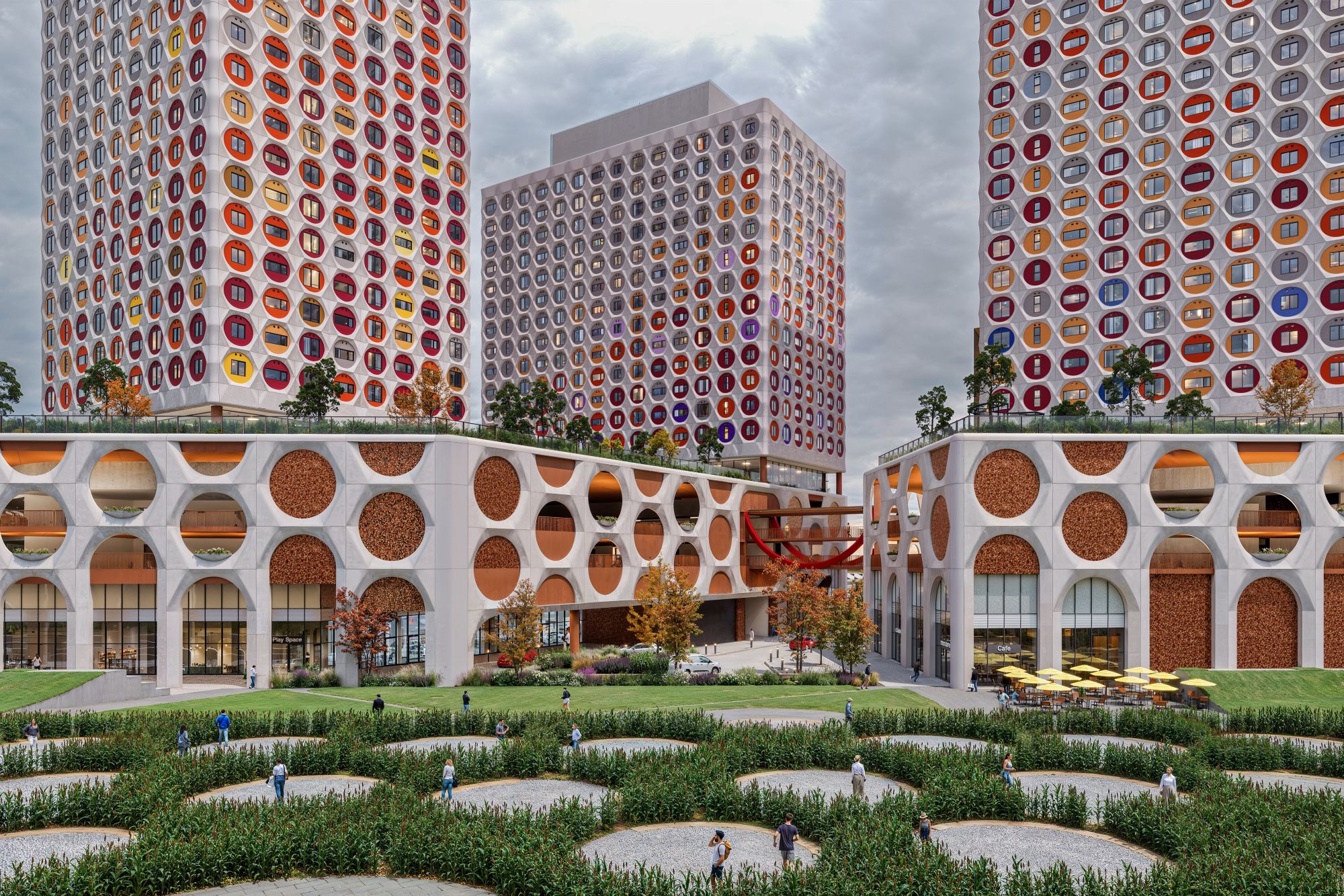 Three colorful, patterned towers rise above arched buildings, landscaped courtyards, winding paths, and people.