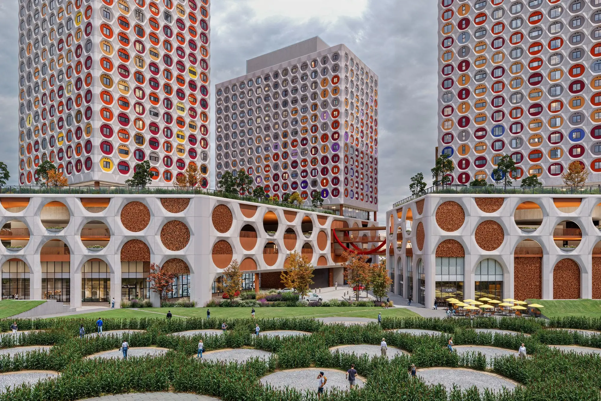 Three colorful, patterned towers rise above arched buildings, landscaped courtyards, winding paths, and people.