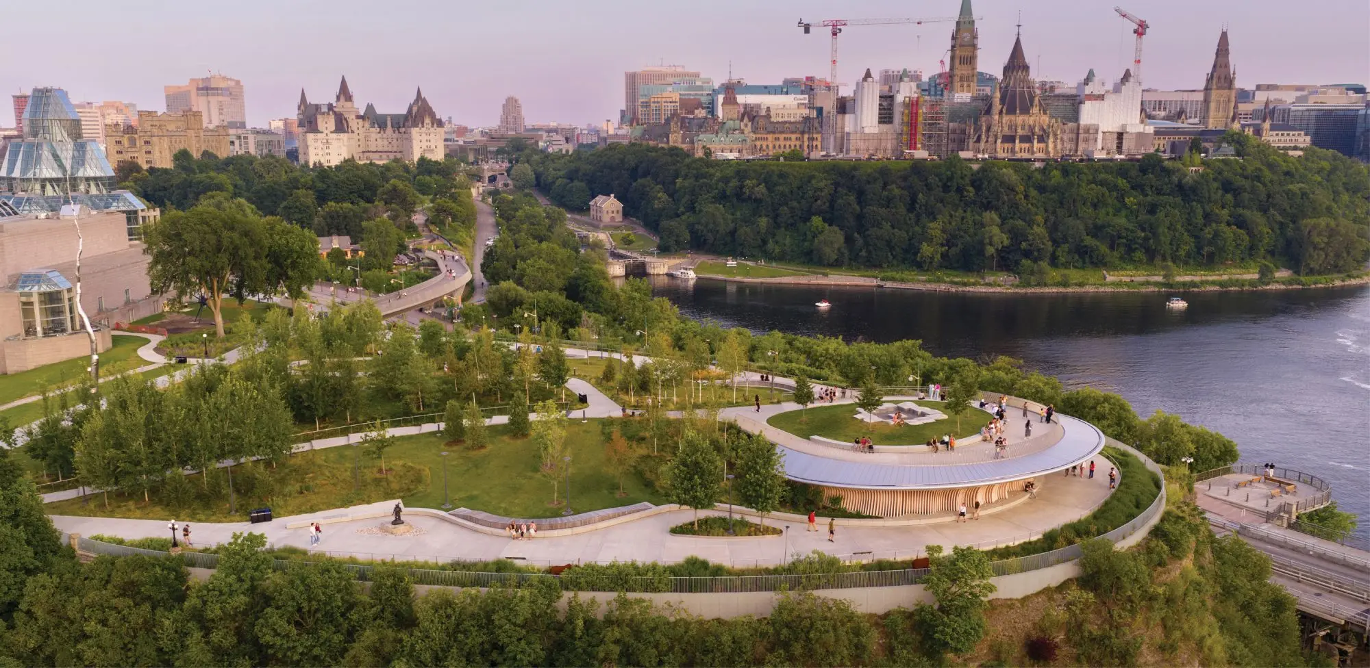 Panoramic view of Ottawa’s waterfront park, river, downtown skyline, and Gothic Parliament buildings.