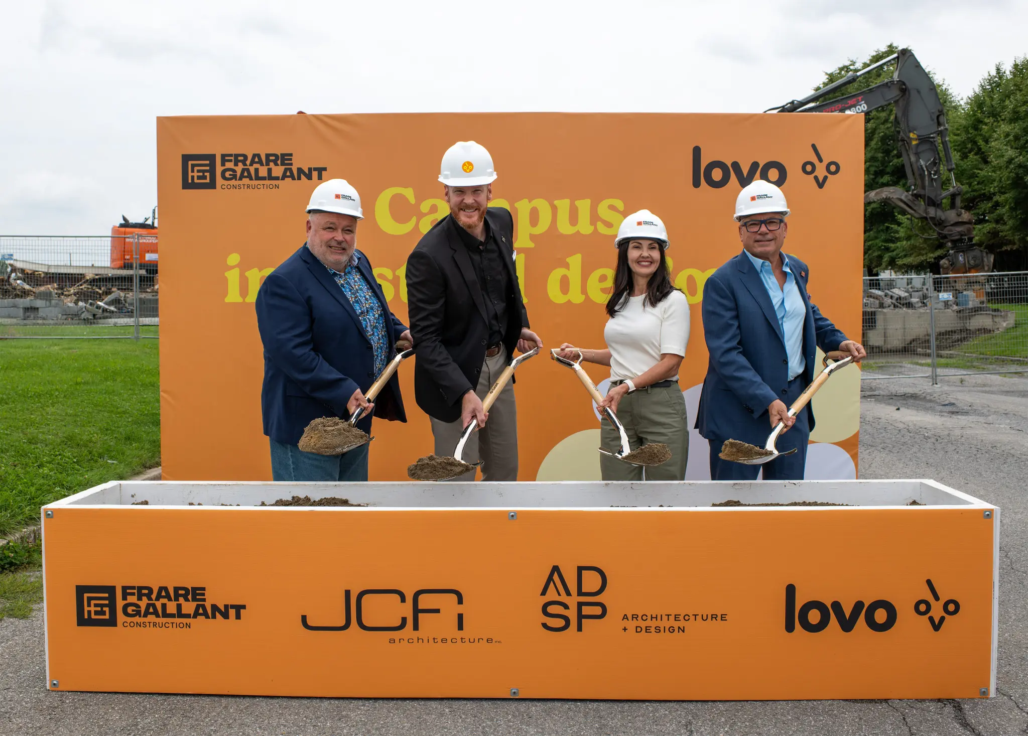 Four adults in hard hats hold soil-filled shovels at an outdoor construction site in front of an orange Campus sign.