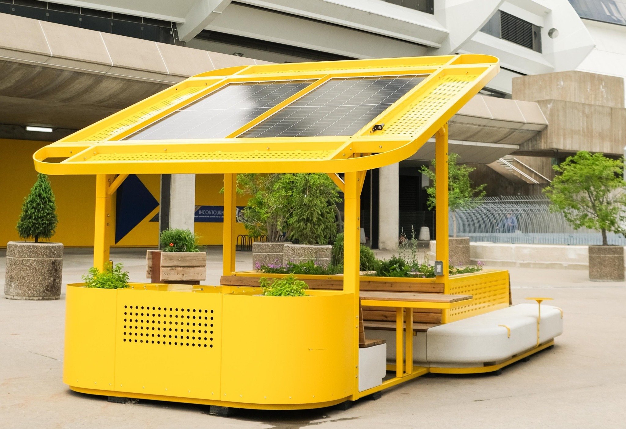 Bright yellow solar-panel-covered outdoor seating and garden structure in a paved plaza.