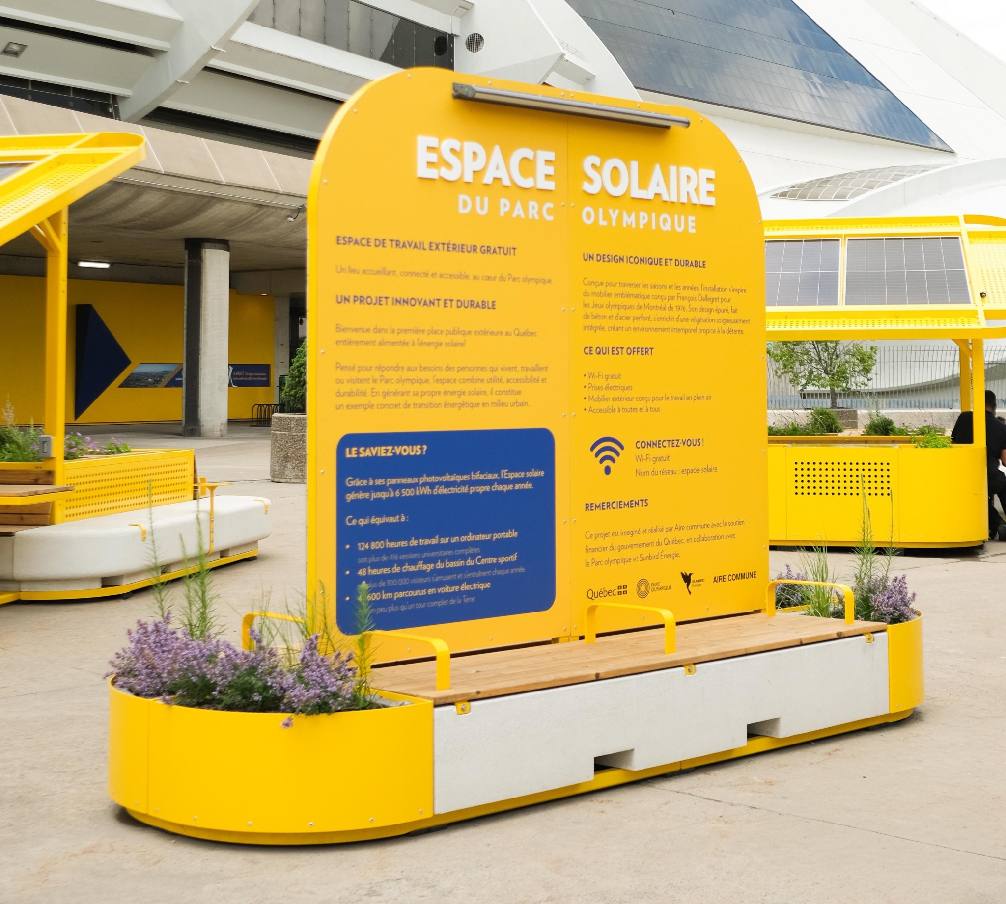 Bright-yellow informational sign for the Olympic Park Solar Space surrounded by outdoor benches and planters.