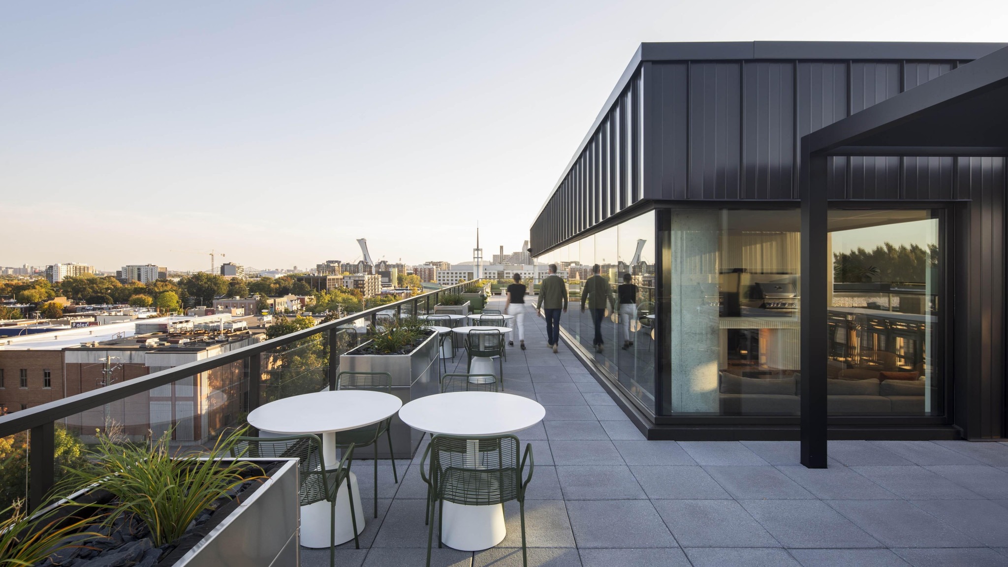 Rooftop terrace with tables, chairs, planters, and people beside a modern building overlooking a city skyline.