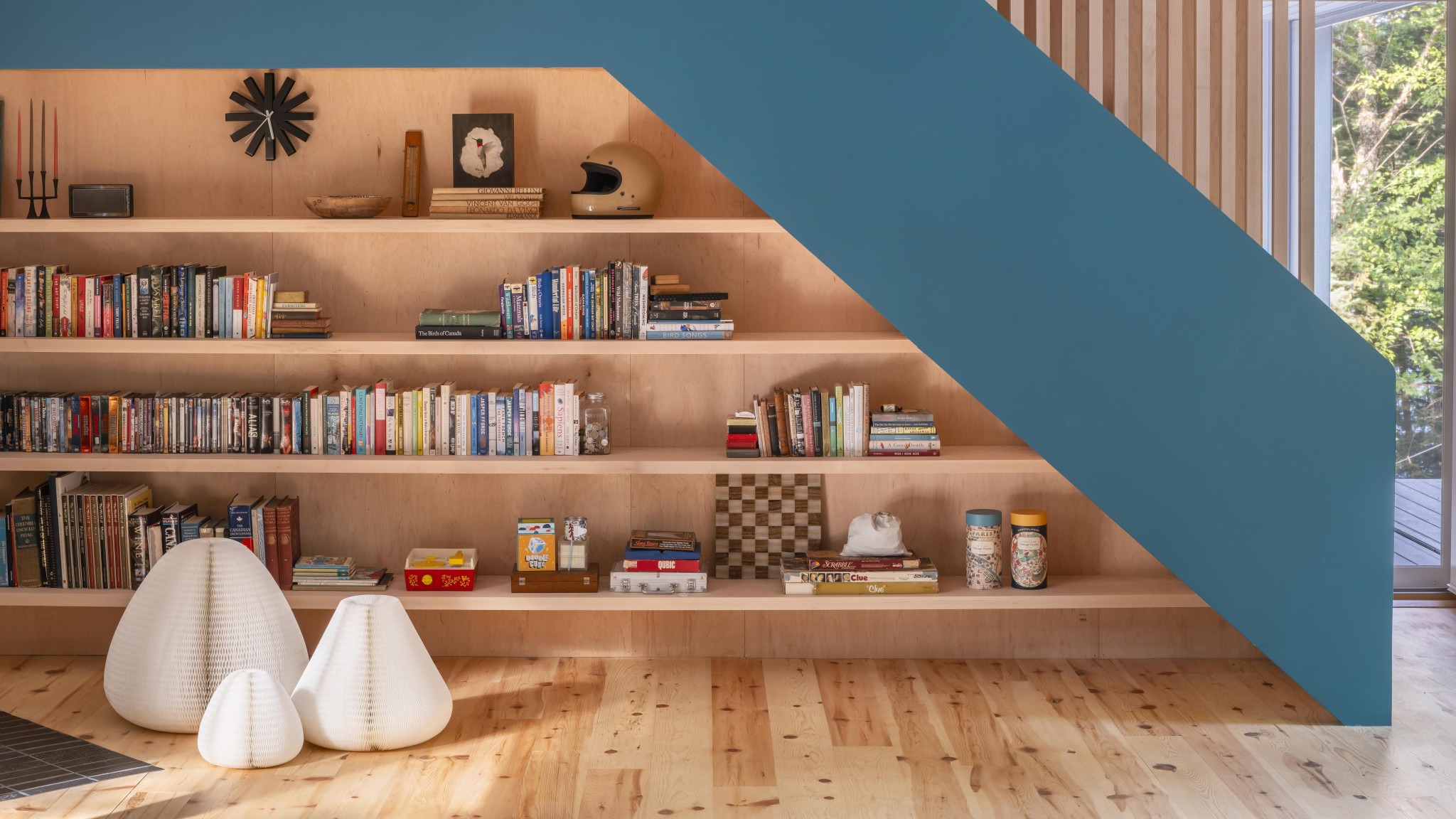 Wooden built-in bookshelves filled with books and games, white sculptural floor lights, and a large blue diagonal beam beside windows overlooking trees.