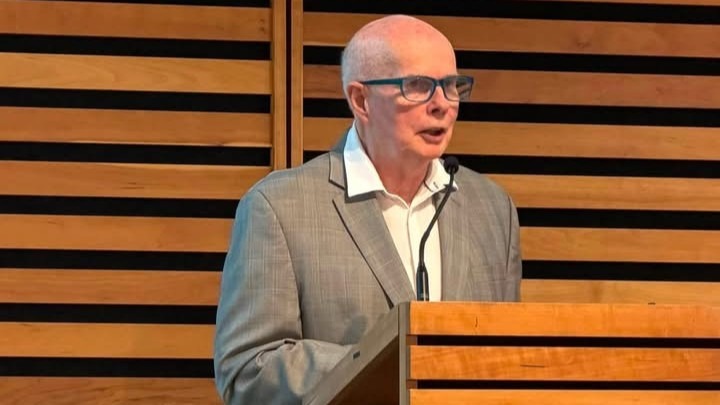 Man speaking at a wooden podium in front of a horizontal slatted wood wall.