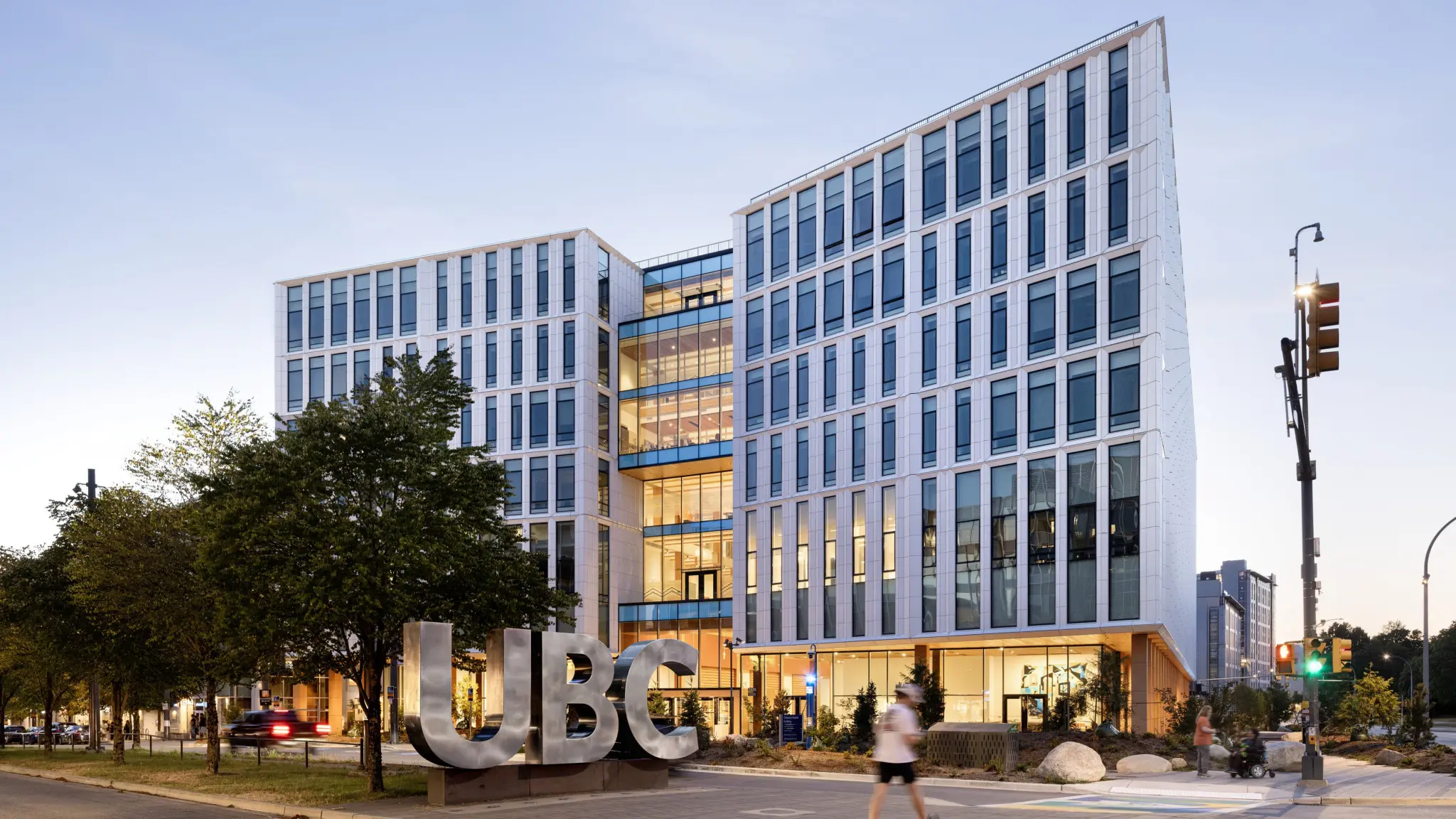 Modern UBC building with a glass atrium, large “UBC” letters, trees, pedestrians, and traffic lights at dusk.