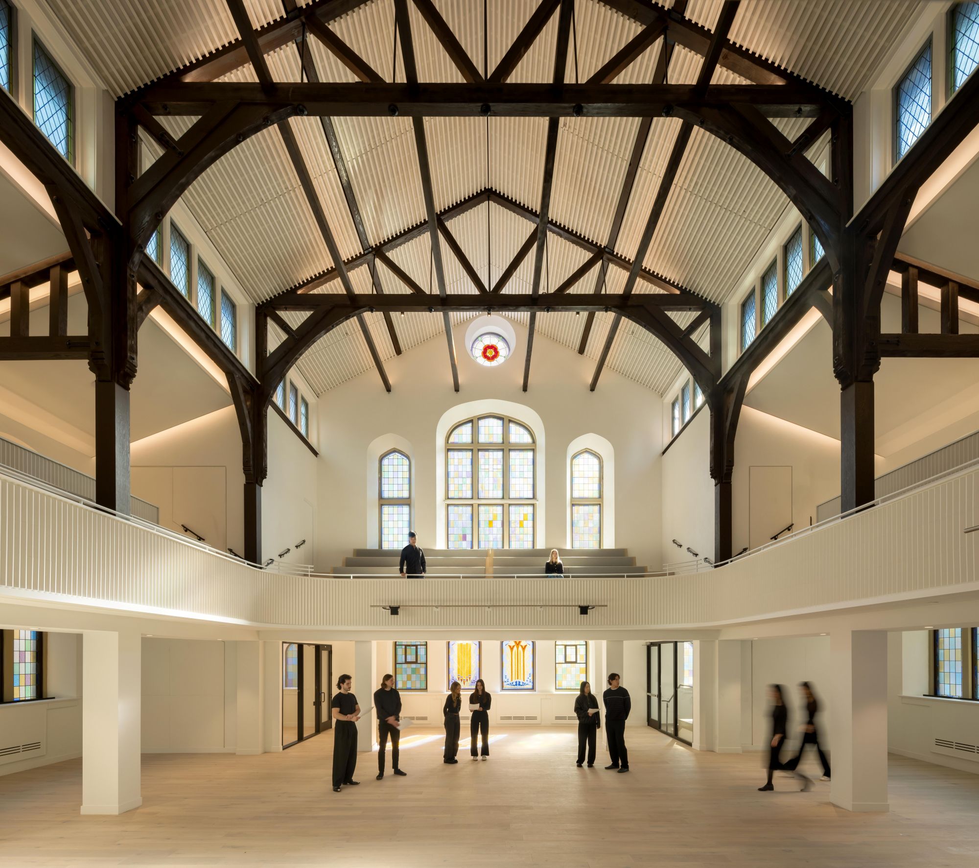 Bright two-story hall with dark wooden roof trusses, stained-glass windows, a balcony, and several people.