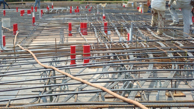 Close view of a reinforcing steel and post-tension cable grid before a concrete pour. Many red cylindrical cast-in firestop devices stand upright among the rebar, with workers visible at the top right.