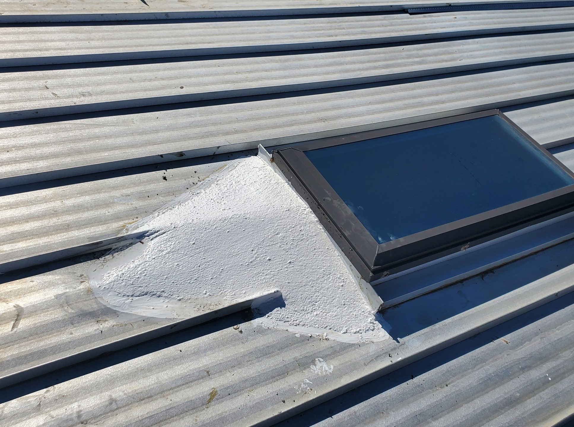 Corrugated metal roof with a skylight in a dark frame. A rough, textured patch of white coating covers the curb flashing and adjacent panel ribs along the skylight’s edge.