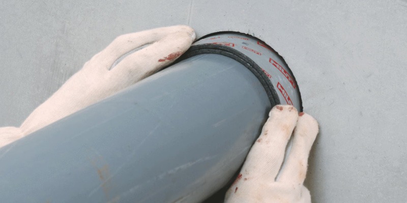 Gloved hands pressing a red-branded firestop wrap strip into the gap around a gray plastic pipe where it passes through a gray wall or floor surface.