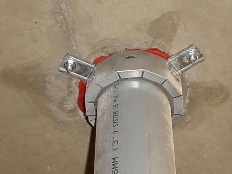 Underside of a concrete slab showing a 3-inch plastic pipe fitted with a metal firestop collar. The collar is anchored to the slab with two steel brackets, and red sealant is visible around the opening.
